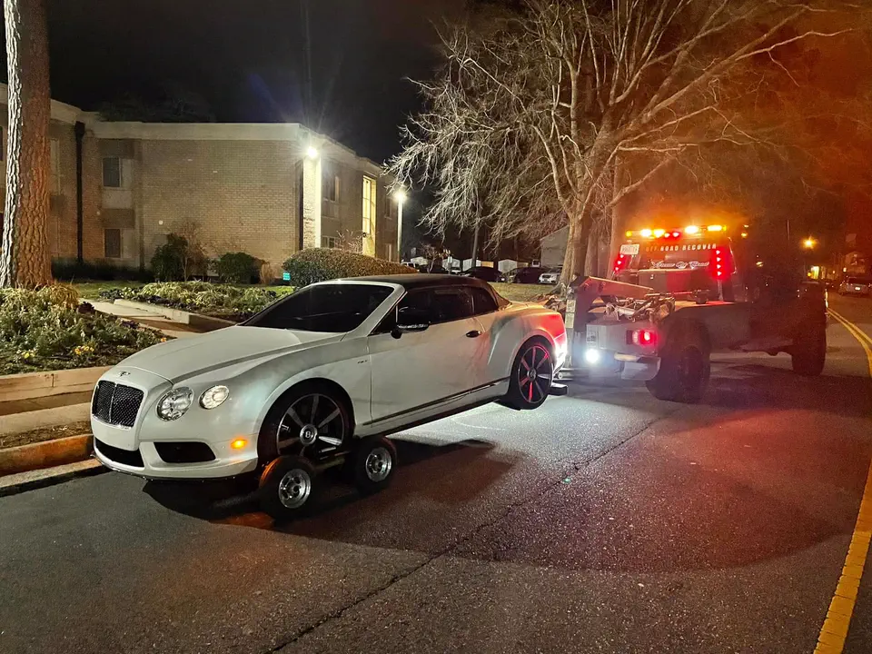 A white Bentley convertible on wheel dollies at night, the tow truck's lights on behind it
