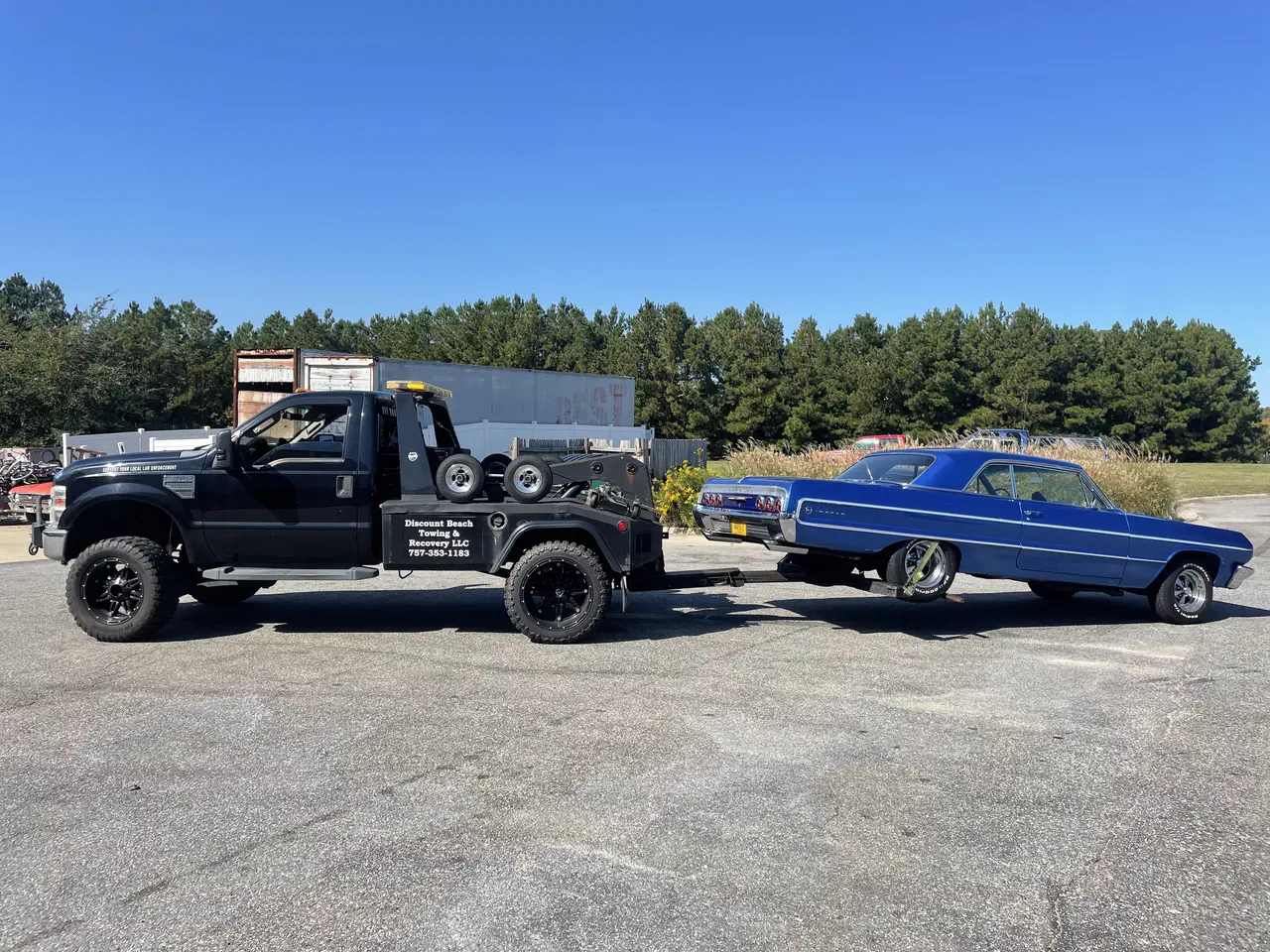 Their black tow truck, lettered Discount Beach Towing and Recovery LLC 757-353-1183 on the side box, with a blue 1960s Chevrolet Impala on the wheel-lift in an open lot