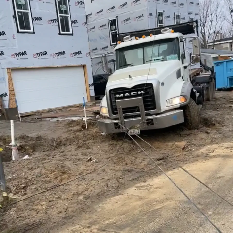 A white Mack dump truck stuck in mud at a building site, with a winch line run out to it