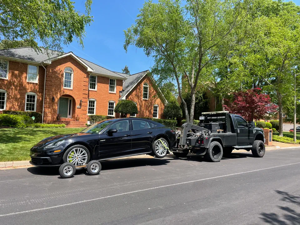 A black Porsche Panamera on wheel dollies behind the tow truck, outside a brick house